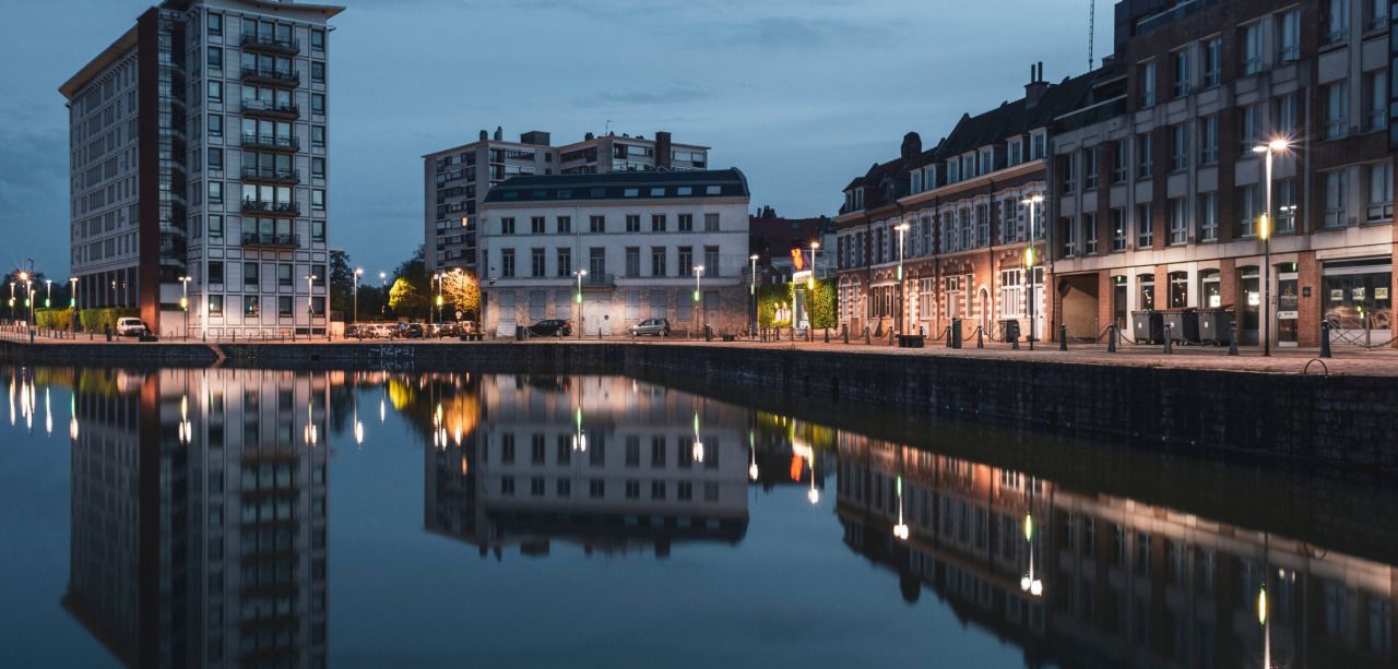 Photographie du quai du wault à Lille, prise au moment de l'heure bleue. Les immeubles ainsi que les éclairages bordant le quai se reflètent dans l'eau en miroir.